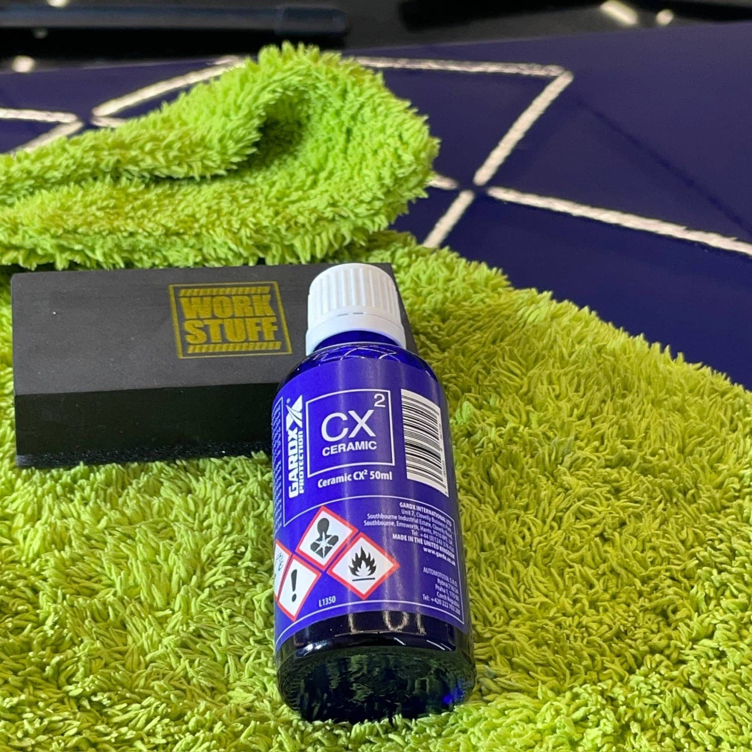 Flaske Gardx Ceramic CX² 50 ml og applikatorblokk på grønn mikrofiberklut, blå lakk i bakgrunnen